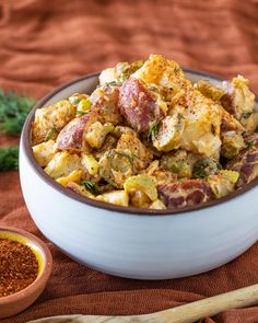 a white bowl filled with potatoes and seasoning next to a wooden spoon on top of a red cloth