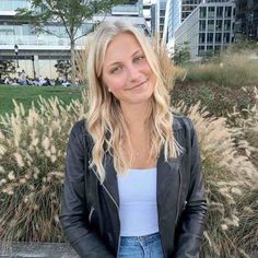 a blonde woman wearing a black leather jacket and blue jeans standing in front of some tall grass