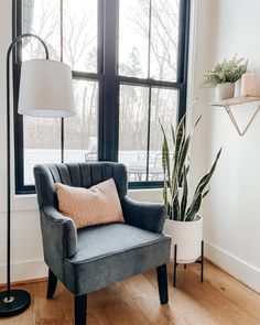 a gray chair sitting in front of a window next to a lamp and a potted plant