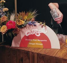 a person pouring orange juice on top of a wooden table next to flowers and drinks