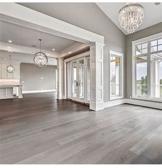 an empty living room with wood floors and chandelier hanging from the ceiling in front of two large windows