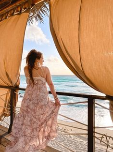 a woman in a pink dress standing on a deck looking out at the ocean and beach