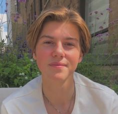 a woman with blonde hair wearing a white shirt in front of some plants and flowers