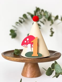 a wooden cake plate topped with a mushroom and number one birthday hat next to greenery