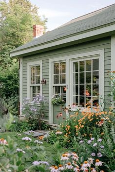 a small green house with lots of flowers in the front yard and windows on each side