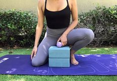 a woman is sitting on a yoga mat with a ball in her hand and an exercise block behind her