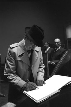 a man in a trench coat writing on a piece of paper with other men behind him