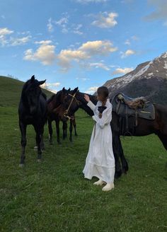 a woman in white dress petting horses on grassy field with mountains in the background