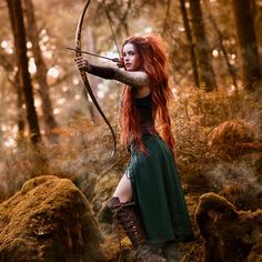 a woman with long red hair is standing in the woods holding a bow and arrow