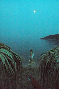 a person standing in the water near some grass and palm trees at night with a full moon behind them