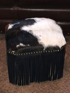 a black and white cowhide ottoman with fringe trim on the bottom, sitting on carpet