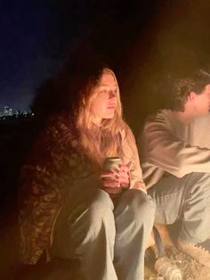 two people sitting next to each other in front of a campfire with the city lights behind them