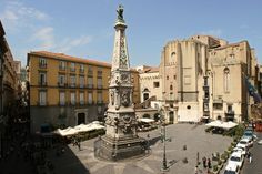 a clock tower in the middle of a city square with people walking around and cars parked on the street