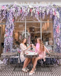 two women sitting at a table in front of a store window with flowers on it