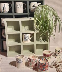 there are many mugs on the table with one plant in front of it and another cup next to it