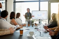 a woman standing in front of a group of people at a table with a projector screen