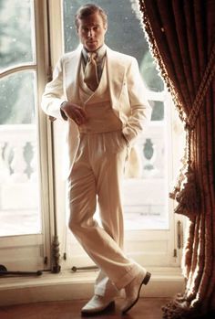 a man standing in front of a window wearing a white suit and tie with his hands in his pockets