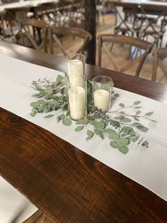 two candles are sitting on a table with greenery in front of them, along with other tables and chairs