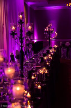 a long table with candles and flowers on it in front of a purple lit room