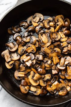 mushrooms cooking in a skillet on top of a marble countertop next to utensils