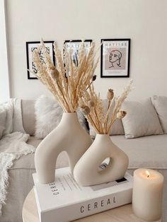 two white vases with dried flowers in them on a table next to a candle