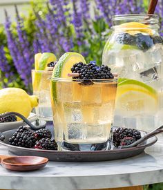 two glasses filled with lemonade and blackberries sit on a marble table in front of lavender flowers