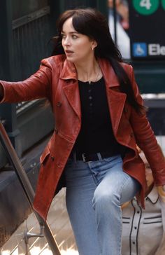 a woman in a red leather jacket and jeans is walking down the street with her luggage