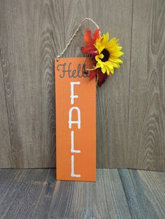 an orange sign with the word fall written on it next to a sunflower and autumn leaves