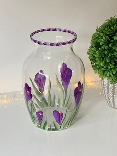 a vase with purple flowers painted on it next to a potted plant and string lights