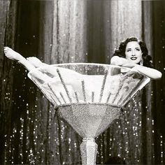 a woman sitting on top of a martini glass in front of curtained backdrops