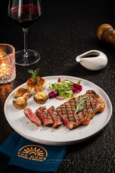 a white plate topped with steak next to a glass of red wine and a candle