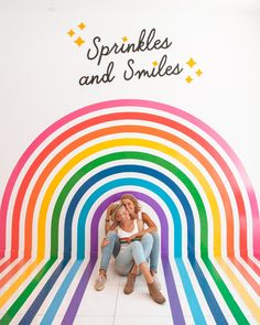 a woman sitting on the ground in front of a rainbow wall with words sprinkles and smiles