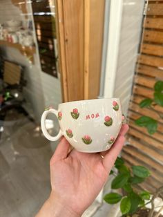 a hand holding a white cup with pink flowers on it and the word mom painted on it