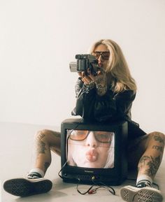 a woman sitting on the floor with her legs crossed and holding a camera up to her face