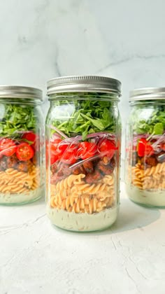 three mason jars filled with pasta salads and veggies on a marble surface