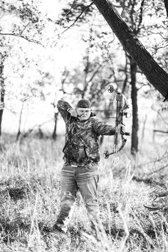 black and white photograph of a man holding a bow in the woods with trees behind him