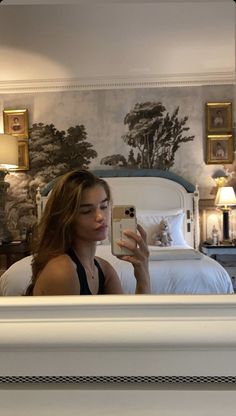 a woman taking a selfie in front of a mirror with a bed and wallpaper behind her