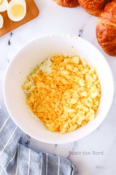 eggs and other ingredients in a white bowl on a marble counter top next to croissants