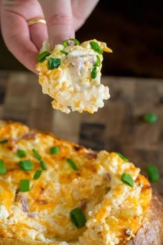 a hand holding a piece of food over a baked potato casserole with cheese and green onions