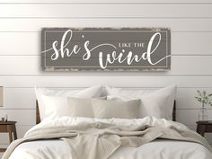 a bed with white sheets and pillows in front of a wooden sign that says she's like the wind