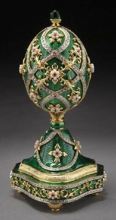 an ornate green and gold vase with flowers on it's base, sitting on a gray surface