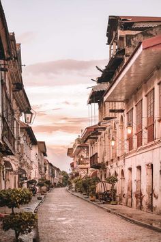 a cobblestone street lined with old buildings and trees in the background at sunset