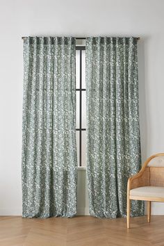 a chair sitting next to a window covered in green floral curtain panels with white flowers on them