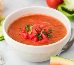 a white bowl filled with tomato soup next to sliced cucumber and watermelon