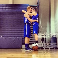 two young men hugging each other in front of a basketball on a court with stairs behind them