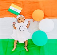 a baby in a white dress and balloons on a green, orange, and white blanket