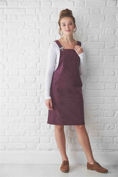 a woman standing in front of a white brick wall wearing an apron dress and brown shoes