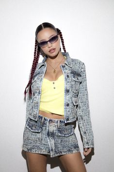 a woman with braids wearing a denim jacket and shorts, standing in front of a white wall