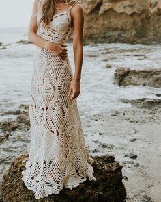 a woman standing on top of a rocky beach next to the ocean wearing a white crocheted dress