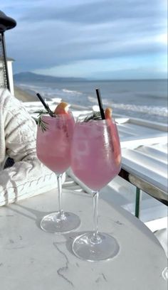 two drinks sitting on top of a white table next to the ocean in front of a woman